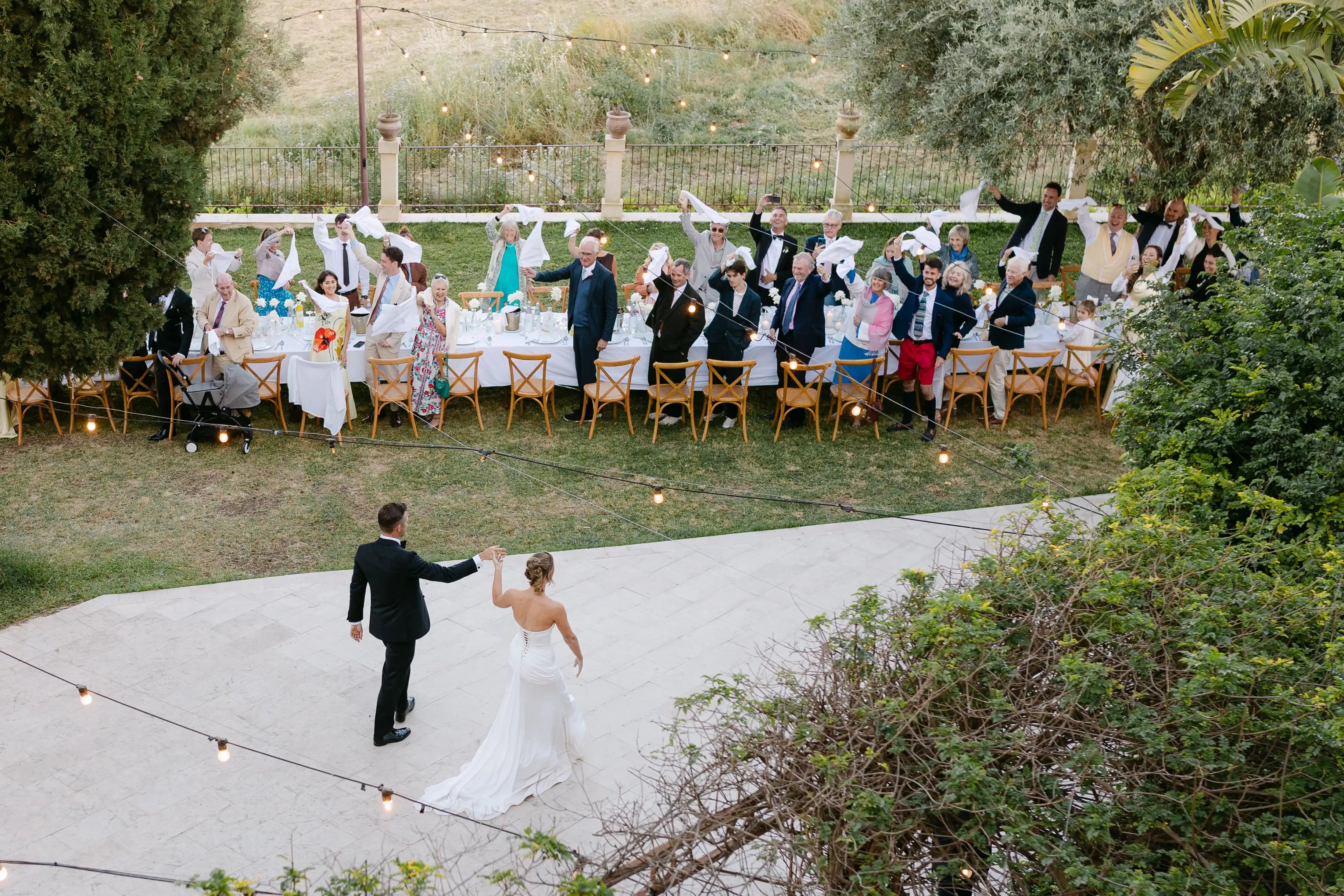 Bride and groom welcomed by guests waving napkins during an outdoor wedding dinner in Italy.